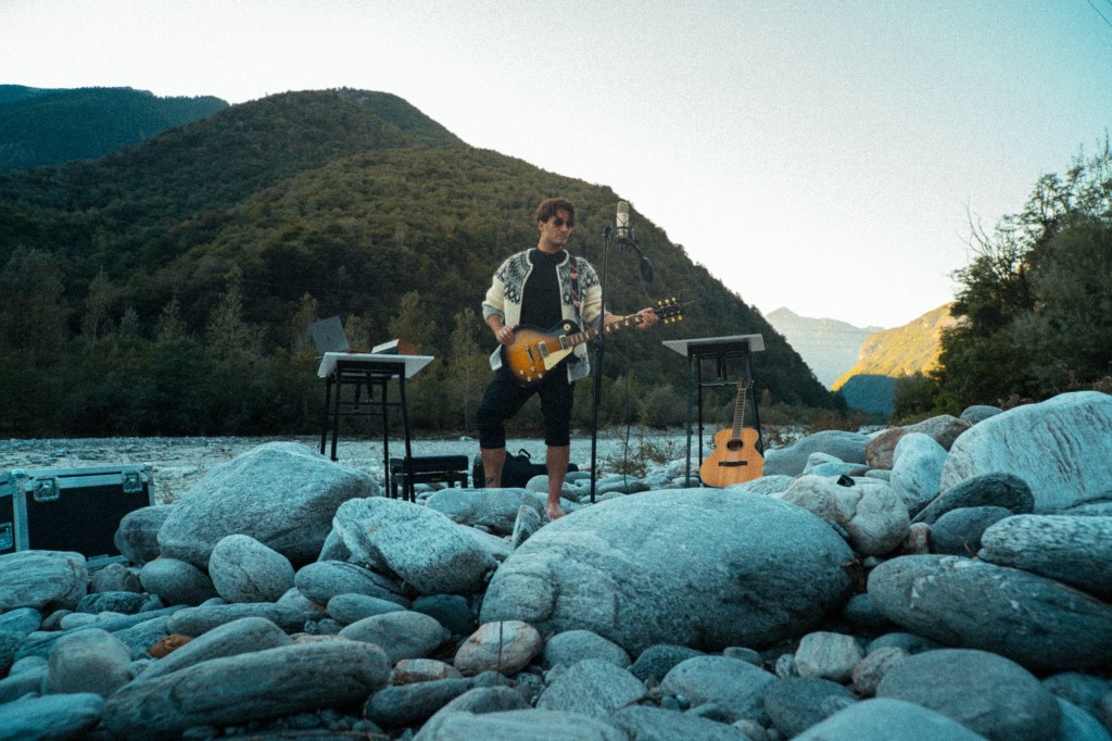 the artist playing electric guitar, with acoustic guitar at its side, in th emountains, surrounded by boulder, green hills in the background