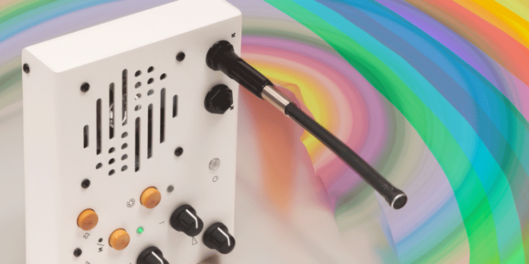 Portable recorder/looper on its side with a rainbow background behind it. White with knobs and wooden buttons, a large speaker grill, and a gooseneck mic protruding perpendicular to the front panel.