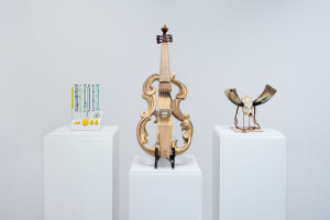 Three speculative designs on white pedestals with a white background. A quipu-powered drum machine with knotted pieces of rope, a viola da gamba with a hollow body and amplification, and a skull-and-bone horn.