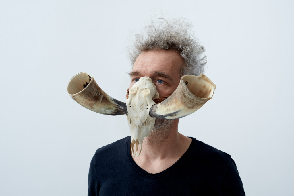 A man wears a skull over his nose and mouth, with two protruding bone skulls out of the sides of the skull - the Craniovox instrument.