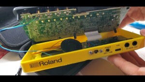 The yellow plastic case of a Roland P-6 pocket drum machine opened up, revealing the factory PCB board and mods with speaker and amplifier circuit.