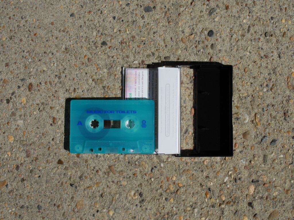 Music for Toilets cassette tape in translucent dolphin blue, against a stone pavement