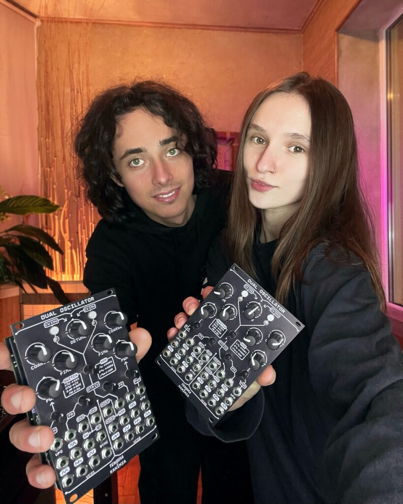 Kaximia and Yehor, two-person Ukrainian Eurorack team of Tembra Modular, smile at the camera holding up dual oscillator modules with a bunch of knobs and CV ints/outs.