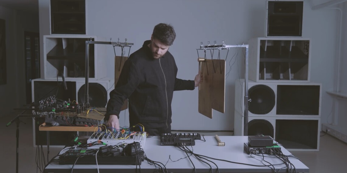 Lorenzo Colombo in live session, framed by speaker stacks, equipment from Torso and Soma on the table and metal percussive plates suspended behind him and on tables, as he strikes one plate with a hammer.