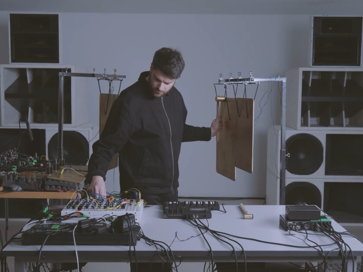 Lorenzo Colombo in live session, framed by speaker stacks, equipment from Torso and Soma on the table and metal percussive plates suspended behind him and on tables, as he strikes one plate with a hammer.
