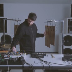 Lorenzo Colombo in live session, framed by speaker stacks, equipment from Torso and Soma on the table and metal percussive plates suspended behind him and on tables, as he strikes one plate with a hammer.