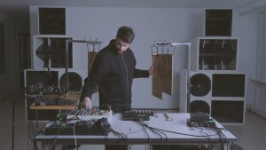 Lorenzo Colombo in live session, framed by speaker stacks, equipment from Torso and Soma on the table and metal percussive plates suspended behind him and on tables, as he strikes one plate with a hammer.