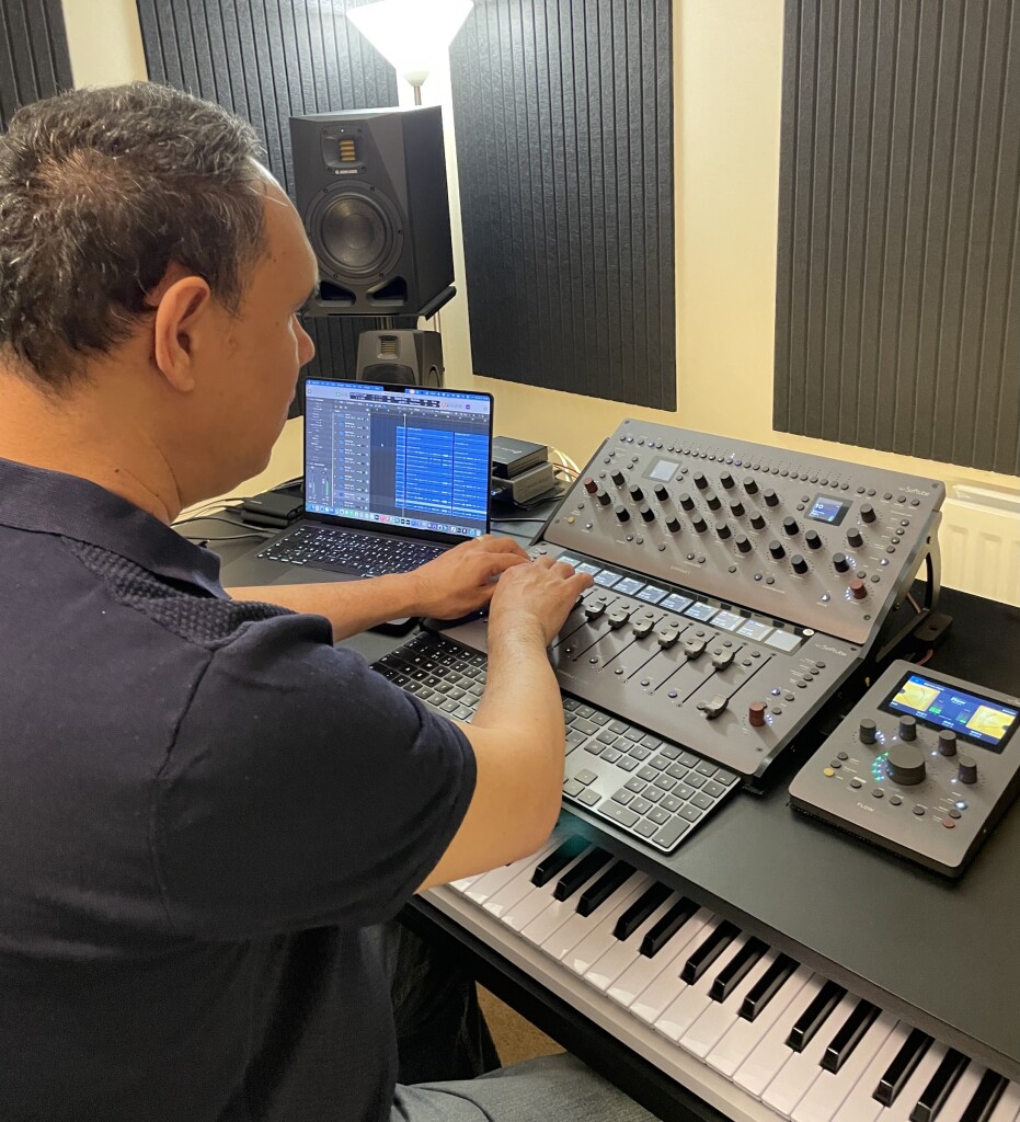 Jason in the studio, viewed from behind, works at a setup with a laptop running Logic Pro and a large Softube Console 1-based digital console controller, his hands on the faders.
