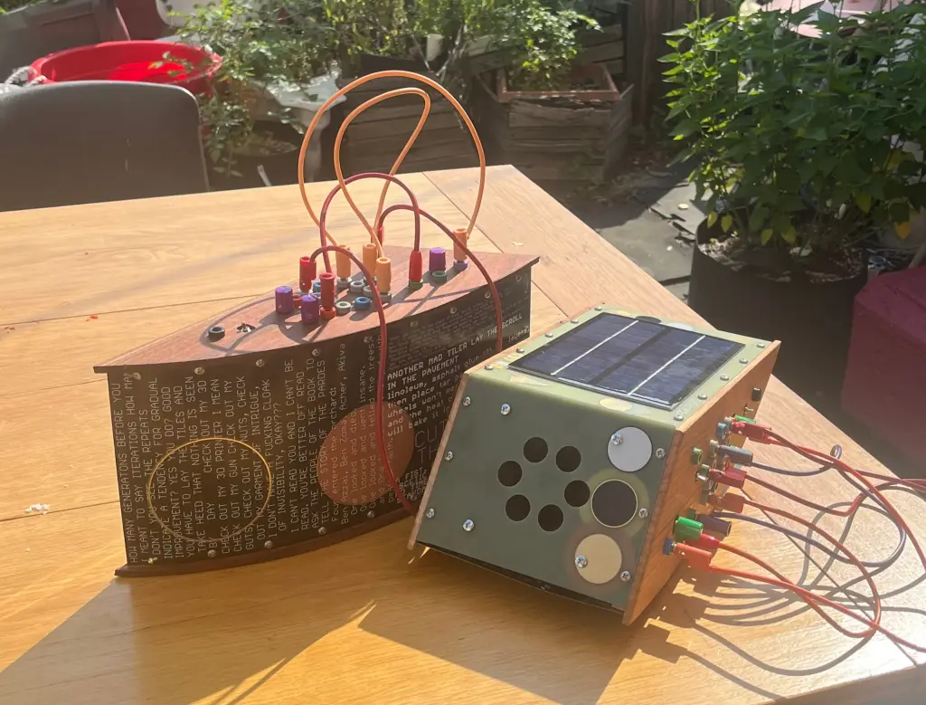 Two Forever Grouse synths sit on a table outside, one with a solar panel facing the sky, another with long inscriptions on circuit board material, wooden side panels, patch cords.